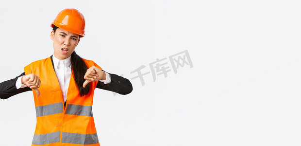 失望和不安的亚洲女总工程师检查糟糕的工作，表现出大拇指向下的失望，戴上安全头盔，抱怨建筑师的糟糕工作，白人背景