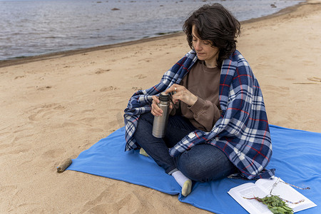 书的海洋摄影照片_活跃的女人喝着热水瓶里的热茶，在海边读书