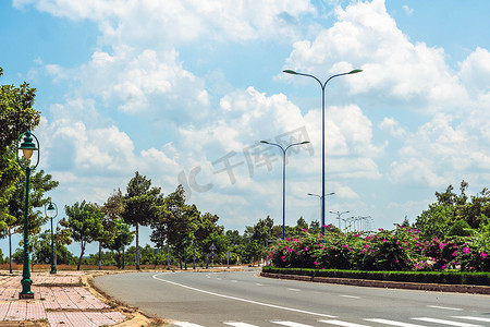 沥青曲线空旷的高速公路，作为分隔线的花丛，白线标记，灯杆灯笼，人行道上的绿树，蓝天积云太阳日。