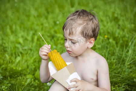 夏天，一个可爱的金发男孩坐在河边多汁的草地上，开胃地吃着玉米。