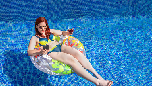 年轻的红发女孩，看着她的智能手机漂浮在泳池内的环形浮标上，同时拿着软饮料。