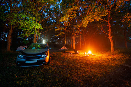 便携式椅子摄影照片_银色汽车附近森林里的夜间篝火和两个帐篷和便携式椅子