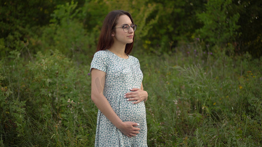 一位年轻的孕妇站着，用手撑着肚子。