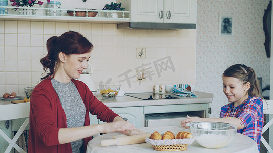 可爱聊天摄影照片_周末，年轻的妈妈一边在厨房做饭，一边揉面团，和可爱的小女儿聊天。