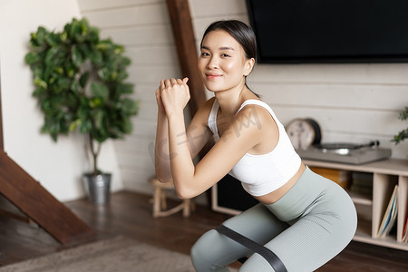 弹力弹力摄影照片_可爱的亚洲健身女孩在家锻炼，腿上拉着弹力绳蹲下，锻炼身体健康