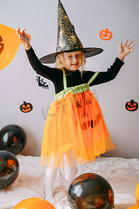 及时通风摄影照片_儿童万圣节-一个戴着女巫帽的女孩和一个狂欢节服装，家里有通风的橙色和黑色气球。