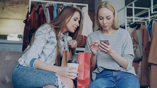 性格开朗的年轻女性坐在漂亮的服装精品店里聊天并使用现代智能手机。