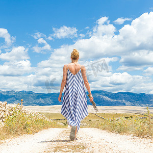 夏日时节，在帕格岛上，穿着夏装的女性手捧薰衣草花束在户外穿过干燥的岩石地中海克罗地亚海岸景观时的后视图