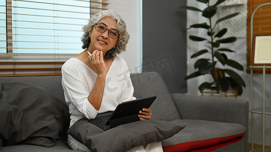 快乐的中年女性在沙发上使用数字平板电脑，在家度过闲暇时光。