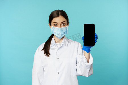 身穿医用面罩和医院制服的年轻女医生，展示手机应用程序、屏幕界面、在线诊所概念，站在绿松石背景上