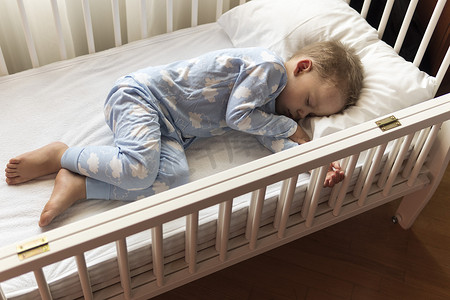 可爱的睡衣摄影照片_顶视图 可爱的小 2-3 岁学龄前男婴在午餐休息时间穿着带枕头的蓝色睡衣在白色婴儿床上甜蜜地睡觉。