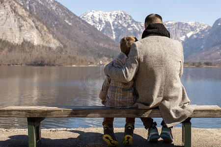 父亲和儿子一起坐在户外的长凳上，享受山、雪、好天气、蓝天。