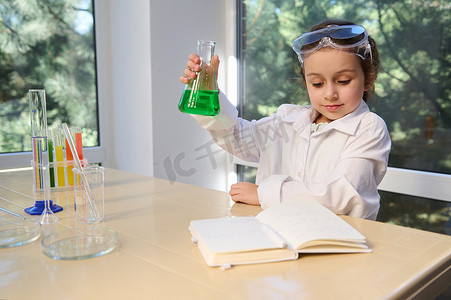 穿着白色实验室外套和安全护目镜的高兴女学生，测试化学实验室实验和摇动玻璃管烧瓶