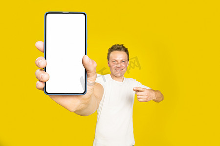 移动应用程序广告英俊的金发男子指着巨大的智能手机与白色的空屏幕，穿着白色 T 恤和牛仔裤隔离在黄色背景。