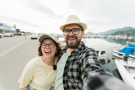 年轻夫妇在户外的夏季港口码头用小船和游艇在智能手机上合影留念时笑着自拍