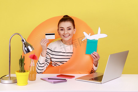 快乐的女人，戴着橡胶圈，展示信用卡和护照，在笔记本电脑上在线订票。