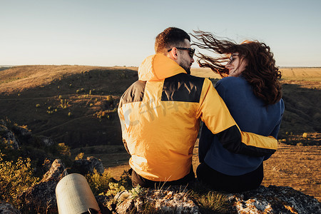 日落时，旅行者夫妇拥抱坐在山边，风吹女人的头发的后视图