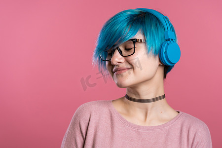 漂亮的年轻女孩，有着绿松石色的头发，在彩色背景下的工作室里玩得很开心，微笑着，戴着蓝色耳机跳舞。