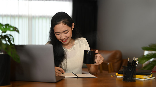 坐在笔记本电脑前的迷人亚洲女性拿着咖啡杯，在笔记本上做笔记