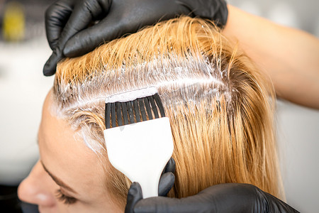 美发师正在美发沙龙里用刷子为一位年轻女子染金色的发根。
