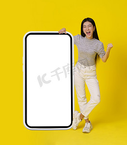 全身的亚洲女孩站在巨大的智能手机旁边，白色的屏幕很高兴，是的，胜利者的手势穿着休闲的黄色背景。