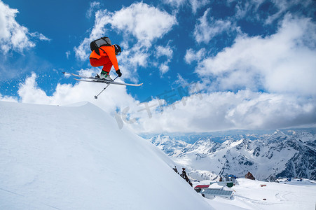一名身着橙色滑雪服的专业滑雪者在蓝天白云的映衬下，从高高的悬崖上纵身一跃，在山间留下一道道雪粉痕迹。