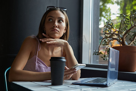 年轻的亚洲美女坐在咖啡馆的桌子旁，拿着笔记本电脑，使用智能手机