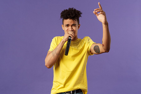 年轻无忧无虑的时髦时髦男人的肖像，纹身，黄色T恤在观众面前表演他的嘻哈或说唱，唱歌，举起手指，拿着麦克风，卡拉OK