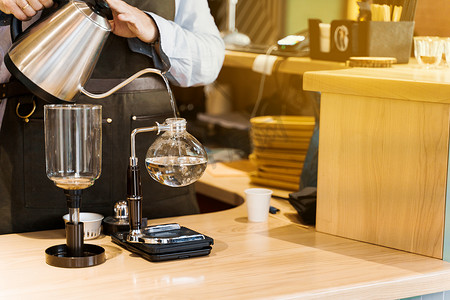 特写咖啡师将水壶中的热开水倒入玻璃虹吸装置中，用于冲泡咖啡。