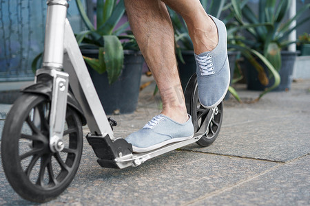 穿着时尚运动鞋的男性脚站在一辆带有大轮子的城镇成人滑板车上，在户外下班后在街道或公园上骑行。