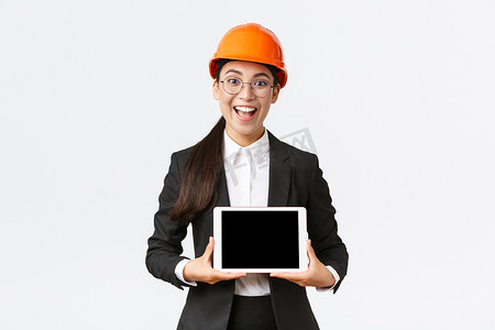 兴奋而惊讶的微笑亚洲女性建筑工程师、安全头盔和西装的建筑师，展示数字平板电脑屏幕，表情惊奇，站在白色背景