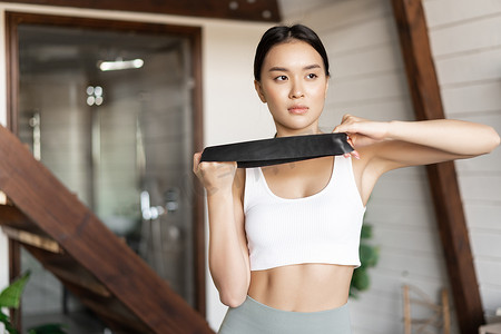 年轻的亚洲女性在家里用弹力绳、手臂二头肌拉伸带、穿着运动服站在客厅里锻炼身体