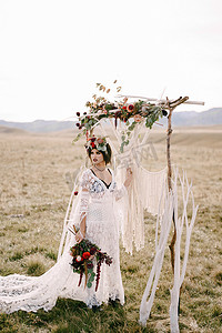 穿着白色柳条裙的新娘和花圈站在婚礼拱门旁边的田野里