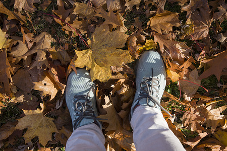 红色黄色枫叶摄影照片_穿着蓝色运动鞋的脚在一堆秋天落下的黄色枫叶中，季节