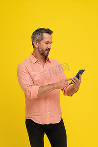 阳刚、健康的中年灰发帅哥拿着智能手机，看着它，穿着黄色的桃色衬衫。