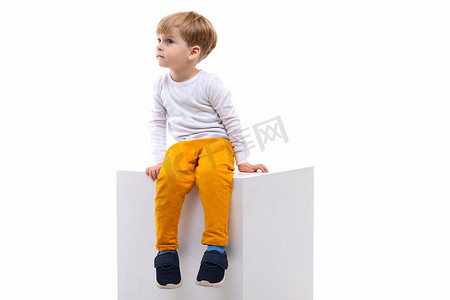 一个好奇的孩子坐在白色立方体上，靠着白墙，向左看