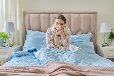 家里生病的年轻女人躺在床上，手里拿着一杯热饮和手帕