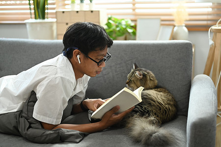 休闲男人和他可爱的猫躺在沙发上看书。