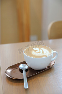 拿杯子摄影照片_咖啡店木桌上的牛奶制成的卡布奇诺或拿铁艺术咖啡