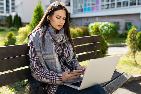 装秋摄影照片_一位身穿秋装的年轻女子坐在外面的长凳上，正专心致志地用笔记本电脑工作