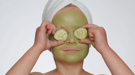 头戴毛巾、微笑的美丽小女孩在脸上涂抹清洁保湿绿色面膜