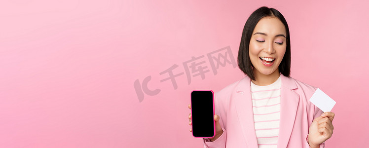 穿着西装、微笑的韩国女商人展示手机屏幕、信用卡、展示网上银行应用程序界面、粉红色背景