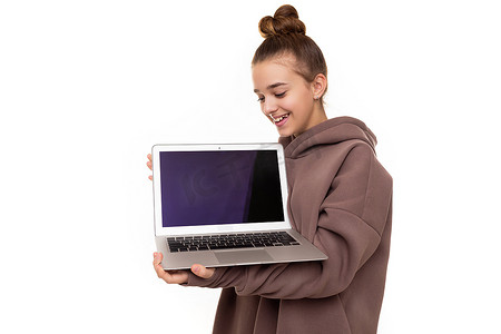 照片中，一个头发黑、头戴棕色连帽衫、带兜帽的漂亮正面可爱女孩拿着一台笔记本电脑，上面有一个空白的黑色屏幕，在白色背景上被隔离，并有可用的文本空间