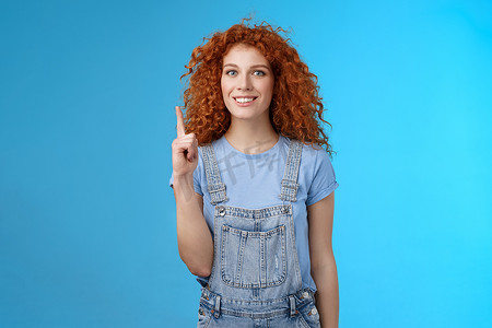欢快可爱、精力充沛的红头发卷发女孩穿着牛仔工作服书暑假旅行优惠好优惠链接指向竖起的食指向上显示广告傻笑