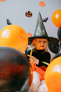 儿童万圣节 — 一个女孩戴着女巫帽，穿着嘉年华服装，家里有通风的橙色和黑色气球。