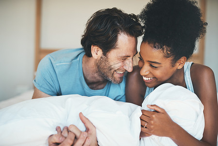 快乐、充满爱和有趣的跨种族夫妇躺在床上微笑着分享温柔的时刻。
