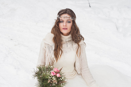 冰雪婚礼摄影照片_美丽的新娘穿着白色连衣裙，在冰雪覆盖的冬季森林里捧着花束。