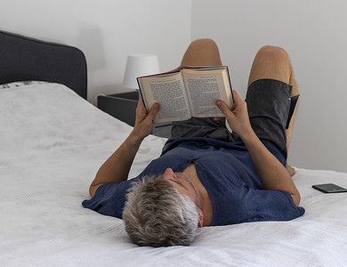 照片中，一名中年男子躺在床上看书。