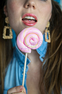 棒棒糖粉色背景摄影照片_蓝色背景、糖果、糖和不健康食品概念上，一个漂亮女孩的肖像，上面有彩色粉色旋转棒棒糖硬糖
