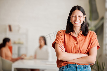 一位自信的西班牙裔年轻女商人双臂交叉站在办公室里，背景是她的同事。